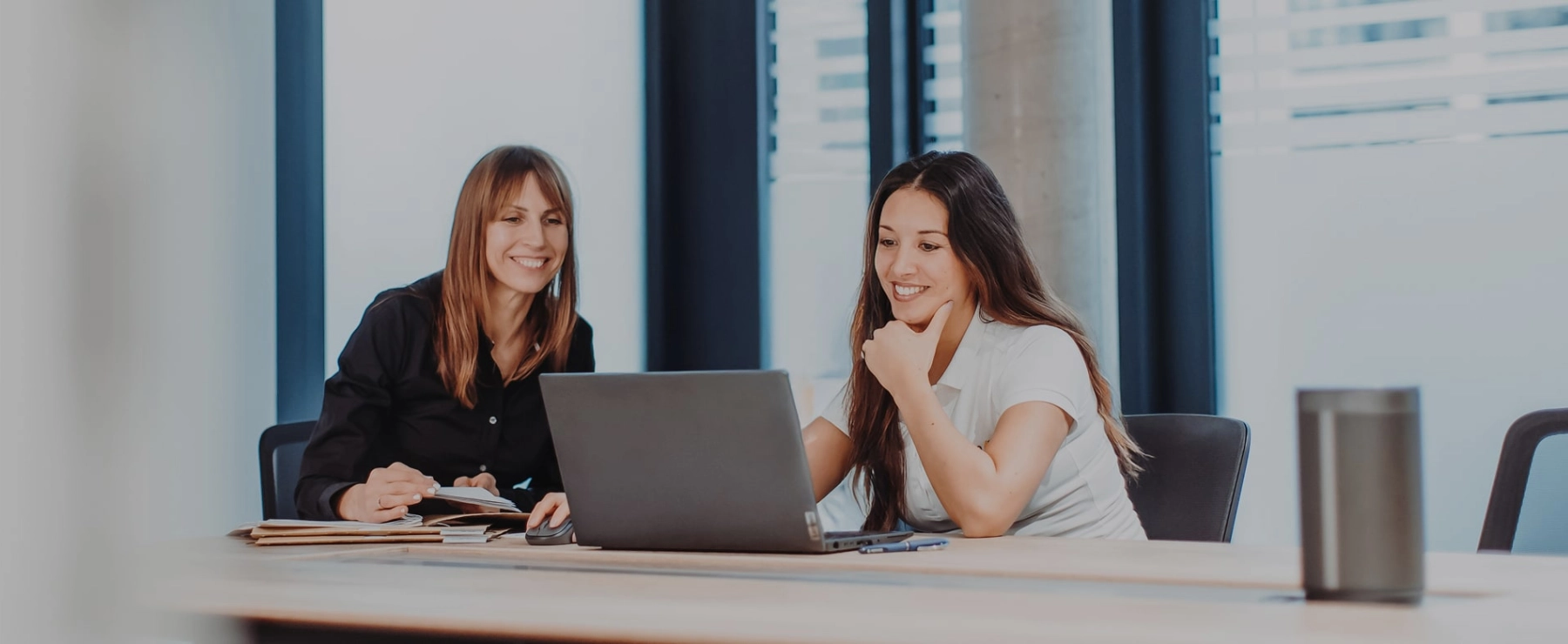Zwei Frauen sitzen an einem großen Bürotisch und lächeln freundlich auf einen Laptopbildschirm