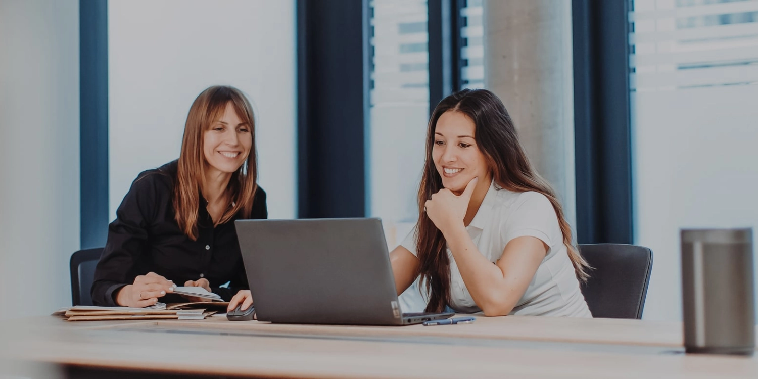Zwei Frauen sitzen an einem großen Bürotisch und lächeln freundlich auf einen Laptopbildschirm