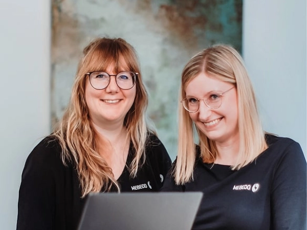 Zwei Frauen stehen gemeinsam in einem Raum, halten einen Laptop und lächeln freundlich